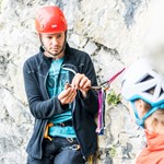 Kletterer mit rotem Helm vor Fels erklärt Person vor ihm Kletterequipment. 
