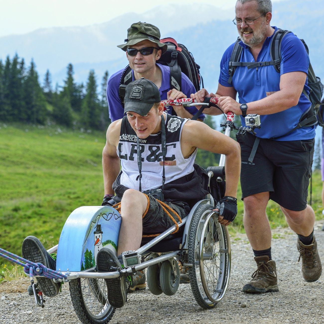 Person im Outdoor-Rollstuhl wird von zwei Personen geschoben, mit Berglandschaft